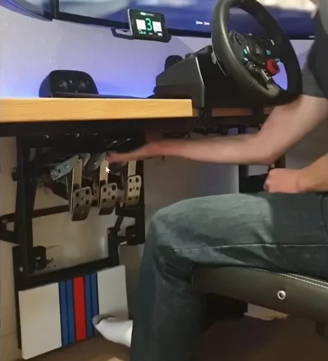 A sim racer seated at a desk with inverted pedals and a heel rest on a tray pulled out from underneath, a cheap sim racing mods answer to a shared desk.