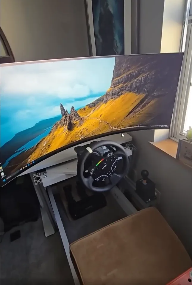 A curved ultrawide monitor slid forward on a rail so it sits directly above the wheel of a desk rig, one of the cheap sim racing mods for shared spaces.