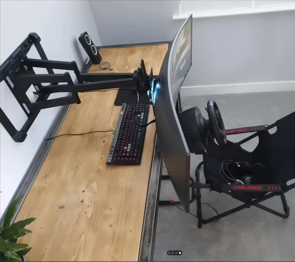An office desk with a wall-mounted monitor arm extended forward over a Playseat Challenge cockpit, one of the cheap sim racing mods for a room that doubles as living space.