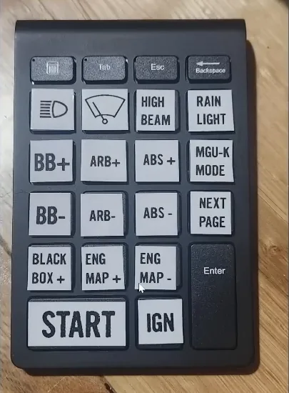 A wireless numeric keypad on a wooden desk with its keys relabelled by printed stickers for brake bias, ABS, engine map, start and ignition, the cheapest of the cheap sim racing mods in the video.