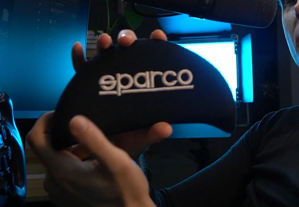 A black Sparco velcro foam seat pad held up in front of the rig, one of the cheap sim racing upgrades that builds a custom support profile on the seat shell.