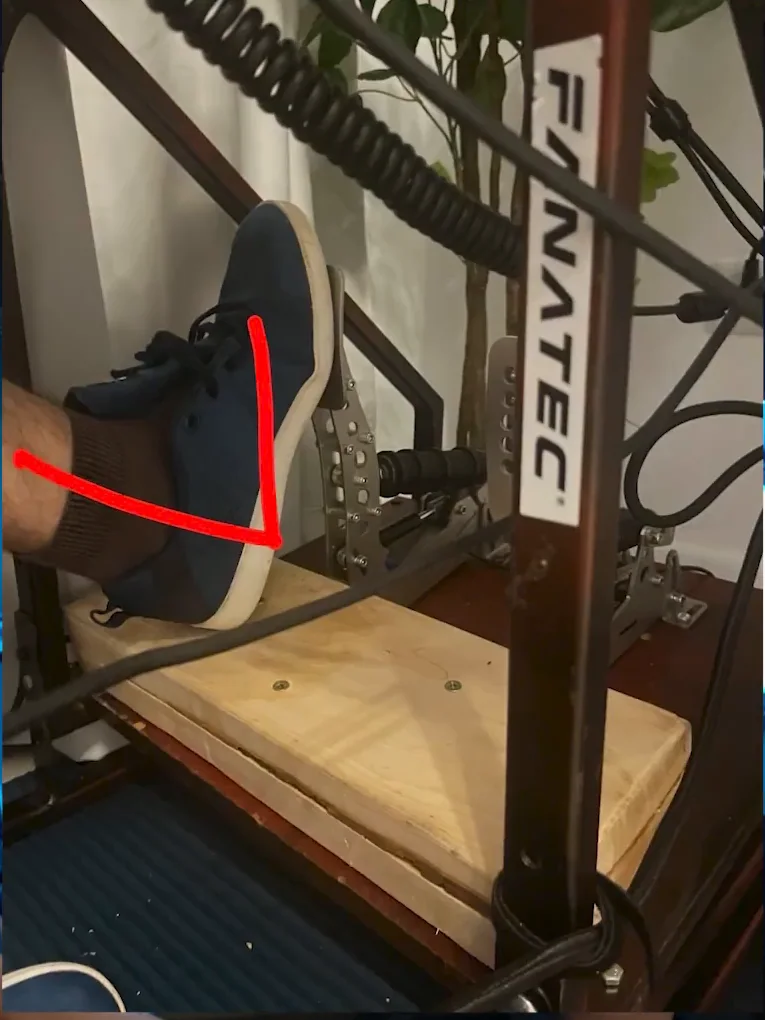 A foot resting on a home-made wooden heel raiser under a pedal set, with a red arrow marking the heel-to-pedal angle, one of the cheap sim racing upgrades you can build yourself.