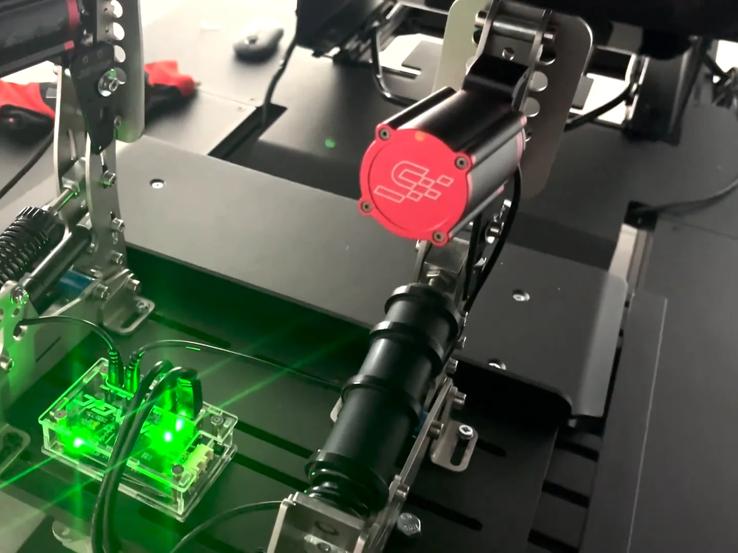 The Simagic haptic control box glowing green on the pedal deck between the pedals, placed close enough for the short reactor cables of a Simagic haptic reactor Heusinkveld mount.