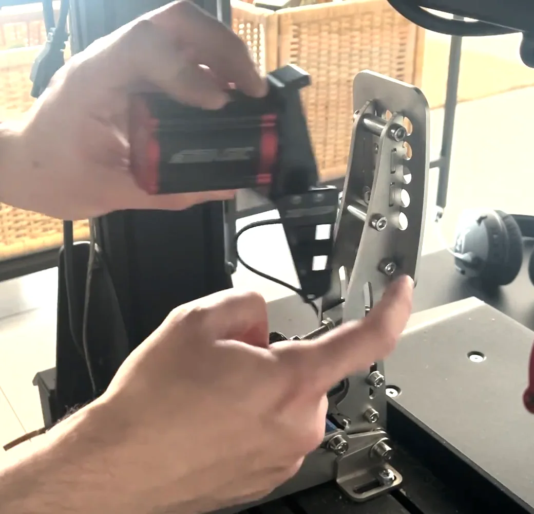 A finger pointing to the existing bolt on a Heusinkveld pedal arm while the assembled reactor is held above it, the bolt reused for the Simagic haptic reactor Heusinkveld mount.