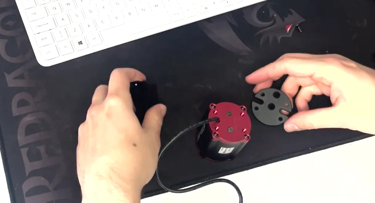 The three parts on a desk before a Simagic haptic reactor Heusinkveld mount begins: the red-capped reactor, the round bracket plate in one hand and the bracket in the other.