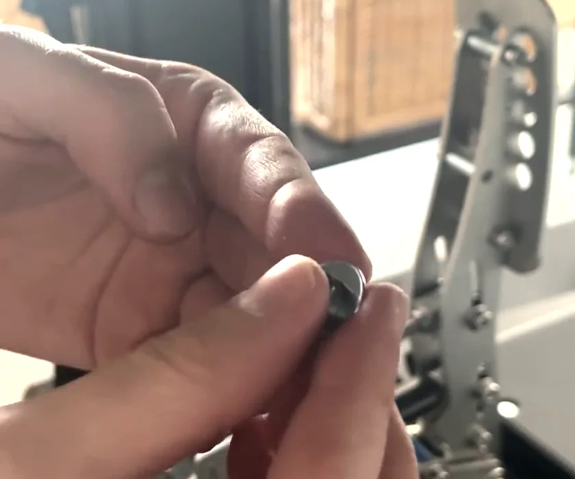 A small washer held between finger and thumb in front of the Heusinkveld pedal, added under the bolt head to tighten the Simagic haptic reactor Heusinkveld mount.